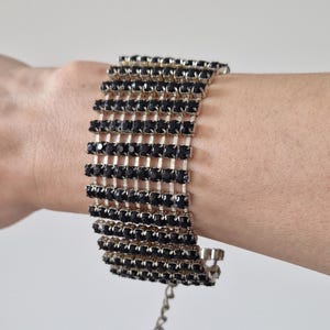 May include: A wide bracelet with multiple rows of black, rectangular rhinestones set in a silver-tone metal. The bracelet is adjustable and has a chain closure. The jewelry is designed to be worn on the wrist.