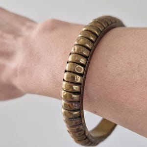 May include: A gold-colored, ridged bangle bracelet. The bracelet is made of metal and has a series of rounded, horizontal ridges. The bracelet is worn on a wrist against a neutral background. Jewelry.
