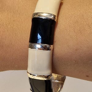 Könnte beinhalten: Ein aufklappbarer Armreif mit abwechselnden, glänzend schwarzen und cremefarbenen Segmenten. Die Segmente sind durch schmale, polierte silberfarbene Metallbänder getrennt, was dem Armreif ein strukturiertes, bambusartiges Aussehen verleiht.
