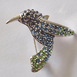 May include: A silver hummingbird brooch, embellished with shimmering blue, green, and purple crystals. The bird's wings are extended, and the brooch has a pin fastening. A lovely piece of jewellery.