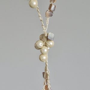 Könnte beinhalten: Cremefarbene Halskette mit einem gedrehten Kordeldesign. Die Halskette hat Cluster aus runden, cremefarbenen Perlen und kleinen, quadratischen, irisierenden Muschelkugeln. Ein zartes, elegantes Accessoire.