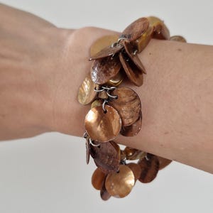 Pode incluir: Uma pulseira com inúmeras contas planas, circulares, em forma de concha, em tons de castanho e dourado. As contas são conectadas por pequenos anéis de metal, criando um efeito pendurado. A pulseira é usada no pulso.