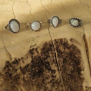 Könnte beinhalten: Vier silberne Ringe mit schillernden Steinen in ovaler, tropfenförmiger und runder Form werden auf einer hellbraunen Holzoberfläche präsentiert. Die Ringe haben aufwendige Metallarbeiten und sind in einer Reihe angeordnet.