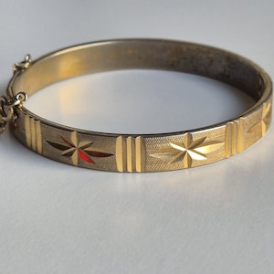 Może przedstawiać: Złota bransoletka typu bangle z zawiasem, posiadająca teksturowaną powierzchnię z powtarzającymi się grawerowanymi wzorami gwiazd i motywami pionowych linii. Bransoletka zawiera mały metalowy mechanizm zapięcia z jednej strony oraz polerowane wykończenie.