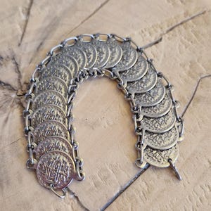 May include: Silver-toned metal bracelet featuring a series of circular, coin-like charms. Each charm is intricately detailed with a pattern. The bracelet is connected by small chain links, and rests on a light brown, textured surface.