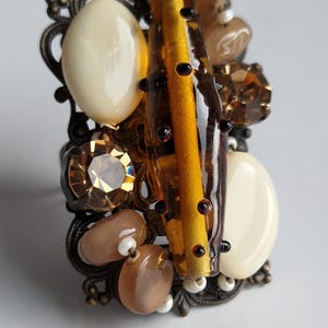 May include: A statement ring featuring an ornate bronze-toned filigree base adorned with a cluster of mixed beads and crystals. The design includes smooth cream-coloured oval cabochons, a central amber-coloured glass rod, and faceted brown crystals.