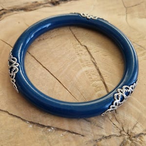 Może przedstawiać: Ciemnoniebieska bransoletka typu bangle ze srebrnymi akcentami filigranowymi. Okrągła bransoletka jest pokazana na drewnianej powierzchni. Srebrne akcenty są równomiernie rozmieszczone wokół niebieskiego paska. Średnica wynosi około 6,5 cm.
