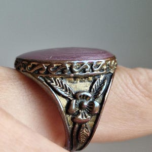 Può includere: Un anello in metallo con una pietra liscia di forma ovale color viola, incastonata in una fascia con finitura ossidata. Il lato della fascia presenta un motivo floreale in rilievo con foglie e un fiore centrale, arricchito da volute.