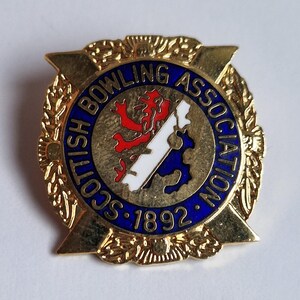 May include: A gold-toned metal pin featuring a circular blue border with the text "SCOTTISH BOWLING ASSOCIATION 1892". The centre displays a red lion rampant and a white diagonal stripe on a textured gold background, framed by four pointed edges.