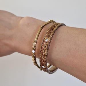 Puede incluir: Tres pulseras doradas en una muñeca. Una es una banda simple con pequeñas piedras transparentes. Las otras dos son texturizadas, con un diseño tejido y piedras transparentes. Las pulseras son de color oro cálido.