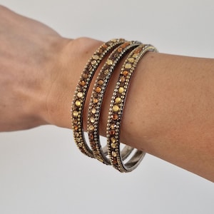Pode incluir: Três pulseiras ornamentadas com base prateada e um padrão de pedras de cor âmbar e dourada. As pulseiras são empilhadas em um pulso contra um fundo liso.