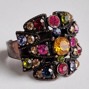 May include: A wide ring with an antiqued copper-toned metal finish featuring an intricate, openwork floral design. The surface is densely set with numerous round, multi-coloured faceted crystals in shades of yellow, pink, blue, green, and amber.
