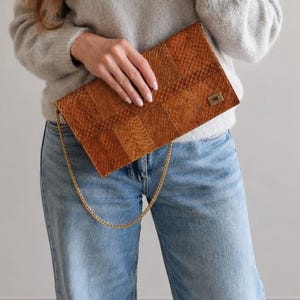 Könnte beinhalten: Eine rechteckige braune Clutch mit geometrischem Muster, einem goldfarbenen Metallverschluss und einem zarten goldfarbenen Kettenriemen. Die Clutch besteht aus einem strukturierten Material, möglicherweise Leder oder lederähnlichem Stoff.