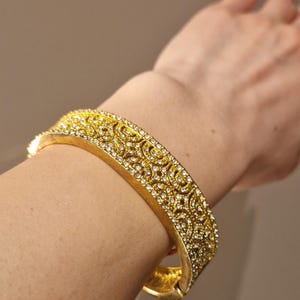 Könnte beinhalten: Ein goldfarbenes Armband mit aufwendigem Filigranmuster und funkelnden, klaren Edelsteinen. Das klappbare Armband wird am Handgelenk getragen und zeigt sein verziertes Design und sein strahlendes Aussehen. Ein Schmuckstück für besondere Anlässe.