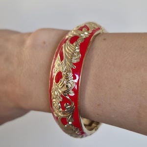 Könnte beinhalten: Ein rotes und goldenes Armreif-Armband mit einem verzierten, wirbelnden floralen Design. Das Armband hat eine glänzende rote Emaille-Oberfläche und goldfarbene Filigranarbeiten. Das Armband hat einen Durchmesser von ca. 6,35 cm.