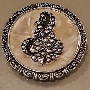 Könnte beinhalten: Runde, silberfarbene Brosche mit floralem Design. Die Brosche hat einen cremefarbenen, perlmuttartigen Mittelteil, umgeben von einem dekorativen silbernen Rand. Das florale Design ist mit kleinen, silberfarbenen Akzenten verziert.
