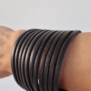 Może przedstawiać: Stos czarnych skórzanych bransoletek otacza nadgarstek. Bransoletki wykonane są z cienkich, płaskich pasków skóry, każdy o szerokości około 0,6 cm. Bransoletki są ułożone blisko siebie, tworząc odważny, teksturowany wygląd.