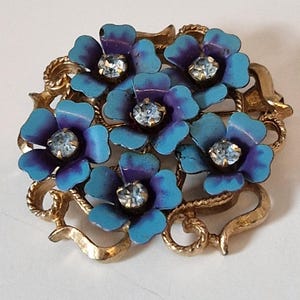 May include: A gold-tone metal brooch featuring a cluster of five flowers with blue and purple enamel petals. Each flower has a clear, faceted rhinestone set in the center. The floral arrangement is framed by a decorative, twisted gold-tone metal border.