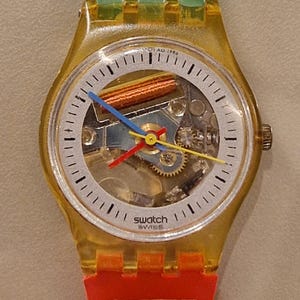 May include: A clear Swatch watch with a transparent yellow case and a colorful dial. The watch band is green at the top with black figures and red at the bottom with black and yellow shapes. The watch face displays the Swatch logo.