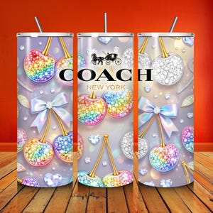 Op de afbeelding: Een cilindervormige beker met een herhalend patroon van regenboogkleurige en witte, met kristallen ingelegde kersen, lichtblauwe strikken en verspreide diamanten op een zachtgrijze achtergrond. De tekst COACH NEW YORK is in het zwart gedrukt.