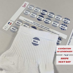 Op de afbeelding: Witte babysok met een cirkelvormig label met de tekst "Alice" in marineblauw. Het label heeft een marineblauw stippenpatroon. Een vel met vergelijkbare labels is zichtbaar op de achtergrond.