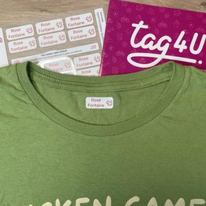 Può includere: Primo piano di una maglietta verde con un'etichetta bianca con la scritta "Rose Fontaine". Sono visibili diversi fogli di etichette bianche con lo stesso testo, insieme a una scatola rosa con la scritta "tag4U".