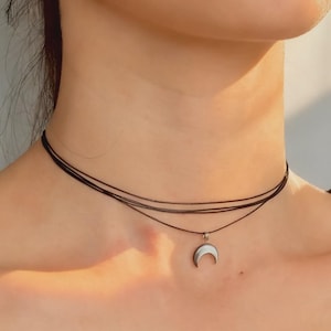 Könnte beinhalten: Eine schwarze Choker-Halskette mit drei Strängen und einem silbernen Halbmond-Anhänger. Der Mondanhänger hat eine weiße, perlmuttartige Einlage. Die Halskette wird um den Hals getragen.
