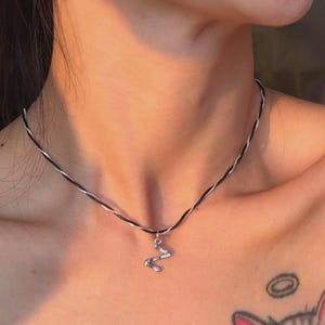 Puede incluir: Collar plateado con un pequeño colgante abstracto, colgado de un cordón blanco y negro. El collar se lleva sobre la piel, con un tatuaje de gato visible en el pecho.