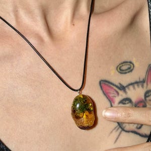 May include: A pendant necklace featuring a smooth, rounded, amber-colored resin stone containing embedded green moss and small yellow elements. The pendant hangs from a simple, thin black cord that rests against the chest area.