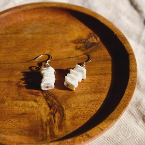 May include: A pair of white shell earrings with silver-colored hooks. The earrings are made of stacked, irregular shell pieces. The earrings are displayed on a wooden plate.