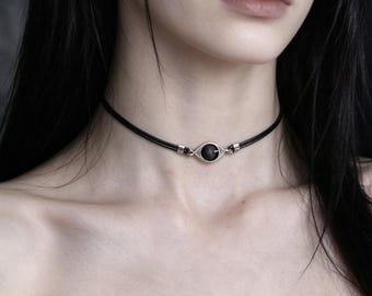 May include: A black cord choker necklace featuring a central silver-toned metal eye-shaped pendant. The pendant contains a small, round, textured black stone at its center, held by silver wire wrapping and connected to the cord by silver metal caps.