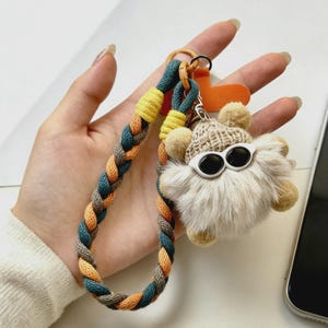 Peut inclure: Un porte-clés fantaisiste avec une créature duveteuse de couleur claire portant des lunettes de soleil et un bonnet en tricot. Le porte-clés a une sangle tressée dans les tons de bleu, orange et gris, avec des accents jaunes. Un accessoire amusant et unique.