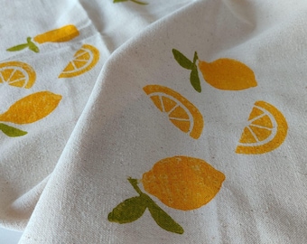 Puede incluir: Primer plano de una tela blanca con un patrón repetido de limones amarillos y rodajas de limón. Los limones tienen hojas verdes. La tela está doblada, mostrando la textura del material.