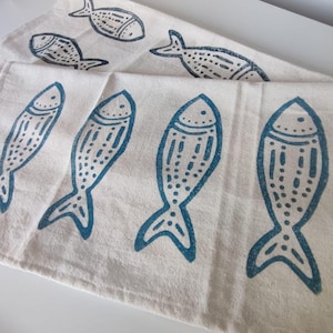 May include: Off-white fabric featuring a repeating pattern of blue fish. The fish are stylized with dotted and lined details. The fabric is folded, displaying multiple fish designs. The overall design is simple and nautical.