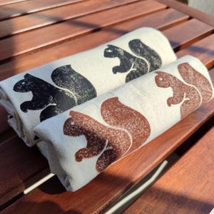 Op de afbeelding: Twee opgerolde beige handdoeken met eekhoornontwerpen in zwart en bruin. De handdoeken zijn gemaakt van een textuurstof en liggen op een houten oppervlak. De eekhoornillustraties zijn eenvoudig en gestileerd.