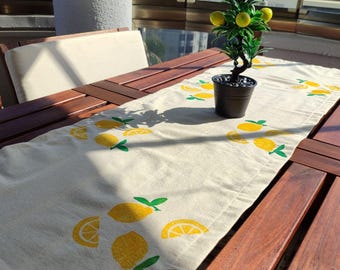 Puede incluir: Un camino de mesa rectangular de color crema con un patrón repetido de limones amarillos y hojas verdes. Un pequeño limonero en maceta se encuentra sobre el camino de mesa, que está colocado sobre una mesa de madera oscura. La escena es en un balcón.