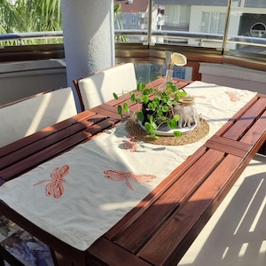 Può includere: Un tavolo da pranzo in legno per esterni con un runner color crema con disegni di libellule arancioni. Il tavolo è apparecchiato con una piccola pianta in vaso, un barattolo di vetro decorativo e un sottobicchiere intrecciato. Sono visibili due sedie bianche imbottite.