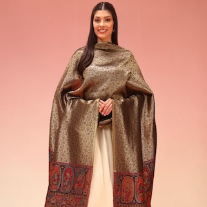 May include: A bronze-colored shawl featuring an intricate floral pattern throughout, with a wide border displaying colorful, detailed illustrations of figures in traditional attire, finished with a fringed edge, worn over a light cream-colored dress.