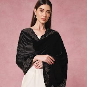 May include: A black velvet shawl with intricate embroidered detailing along the edges. The shawl is draped over a white garment, creating a contrast. The background is a soft pink.