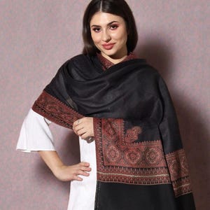 May include: A black shawl with a detailed, reddish-brown border. The shawl is draped over a white garment. The border features an intricate paisley and floral design. The shawl has a fringed edge.
