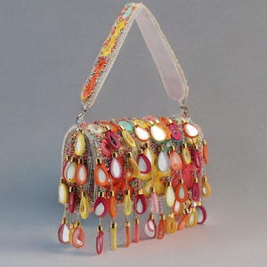 May include: A colourful handbag with a white handle and a rectangular body. The bag is decorated with teardrop-shaped embellishments in orange, yellow, pink, and white. The handle features colourful embroidery. The bag is a statement piece.