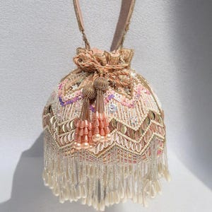 Puede incluir: Un bolso redondo con cuentas en tonos rosa, dorado y crema. El bolso presenta intrincados trabajos de lentejuelas y cuentas, un cierre de cordón y un asa. Largas borlas con cuentas cuelgan de la parte inferior, lo que se suma al diseño ornamentado del bolso.