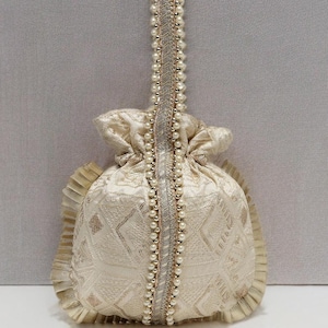 Puede incluir: Una bolsa con cordón de color marfil con un asa decorativa. La bolsa presenta un patrón texturizado y está adornada con perlas y detalles dorados. El asa está adornada con perlas y una cinta plateada.