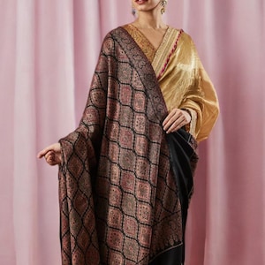 Puede incluir: Una mujer con un chal estampado con borde negro. El chal presenta un diseño geométrico repetitivo en tonos marrón, rojo y verde sobre un fondo negro. Lleva un top dorado y pendientes.