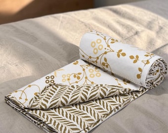 May include: A white cotton blanket featuring a mustard yellow floral and vine print on one side and a mustard yellow leaf pattern on the reverse. The blanket is partially rolled, showcasing both the floral design and the dense leaf print.