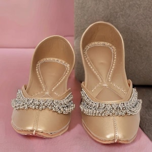 May include: A pair of gold-colored, flat shoes with a rounded toe. The shoes have a decorative beaded trim across the top. The background is a soft pink and gray.