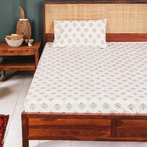 Puede incluir: Una cama con estructura y cabecero de madera, con juego de sábanas y almohada blancas. La ropa de cama tiene un diseño floral repetido en rosa y verde. La habitación tiene una estética cálida y acogedora.