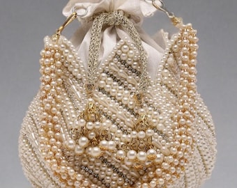Puede incluir: Un bolso tipo bolsa de cuentas de color blanco roto con cierre de cordón. La bolsa está cubierta de perlas y pequeñas cuentas, con detalles y borlas doradas. La bolsa tiene forma redonda y fruncida.