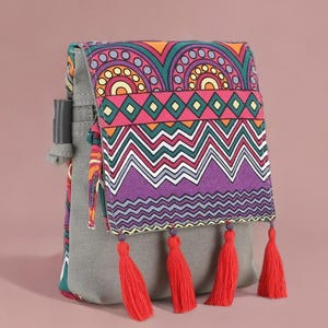 Puede incluir: Una mochila gris con una solapa colorida con patrones geométricos en naranja, morado, verde y blanco. La solapa tiene cuatro borlas rojas. La bolsa tiene un cierre de cordón y una correa gris.
