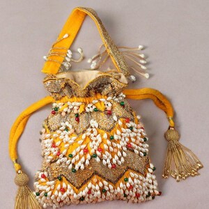 Peut inclure: Un sac jaune à cordon avec une poignée et des glands dorés. Le sac est orné de perles blanches, rouges et vertes, et de broderies dorées. Le motif du sac présente un motif en zigzag.
